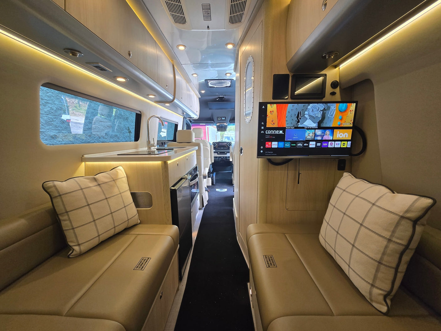 Interior of an rv rental features a compact kitchen with sink, comfy bench seating with pillows, and a wall-mounted TV for entertainment.