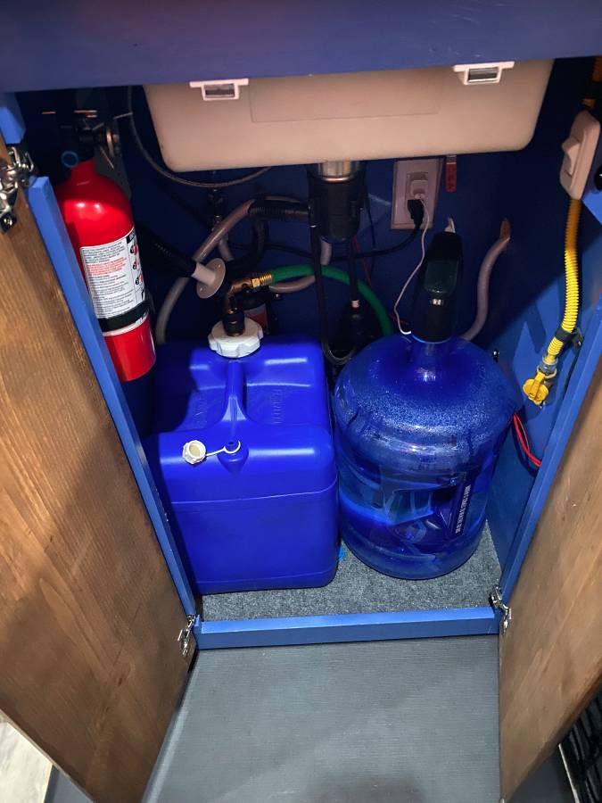 RV rental under-sink utility: water tanks, pump, electrical, plumbing, and a fire extinguisher for safety.
