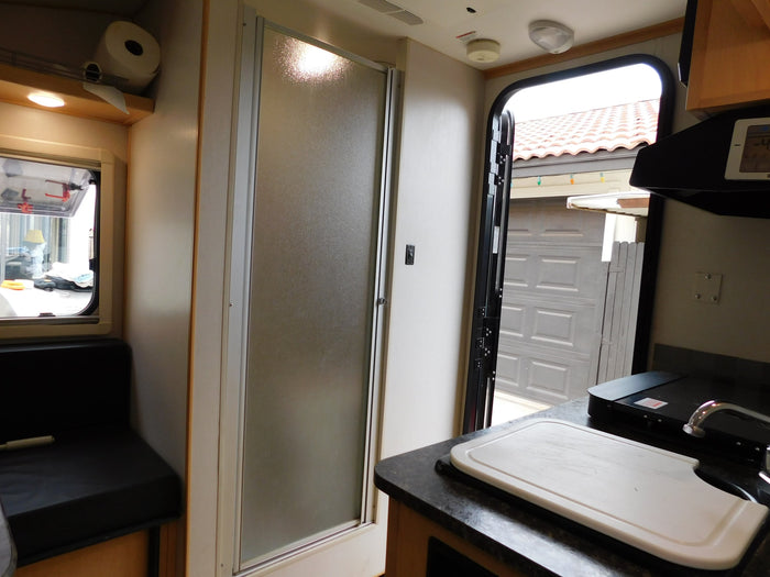 Interior view of an RV rental showing the kitchen with a sink, cutting board, a frosted shower door, and an open entry door.
