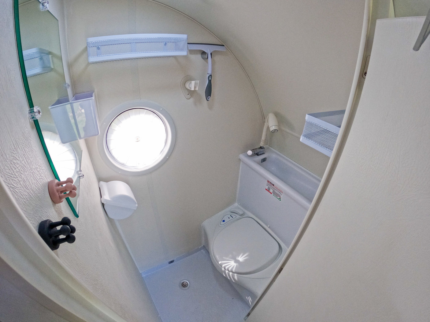 Compact RV rental wet bath with toilet, shower, mirror, and small round window, designed for efficient use of space.