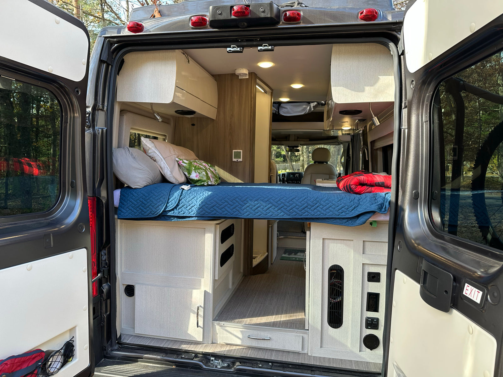 Spacious RV rental interior featuring a cozy bed, cabinetry, and controls, viewed from the open rear doors. Ideal for adventures.