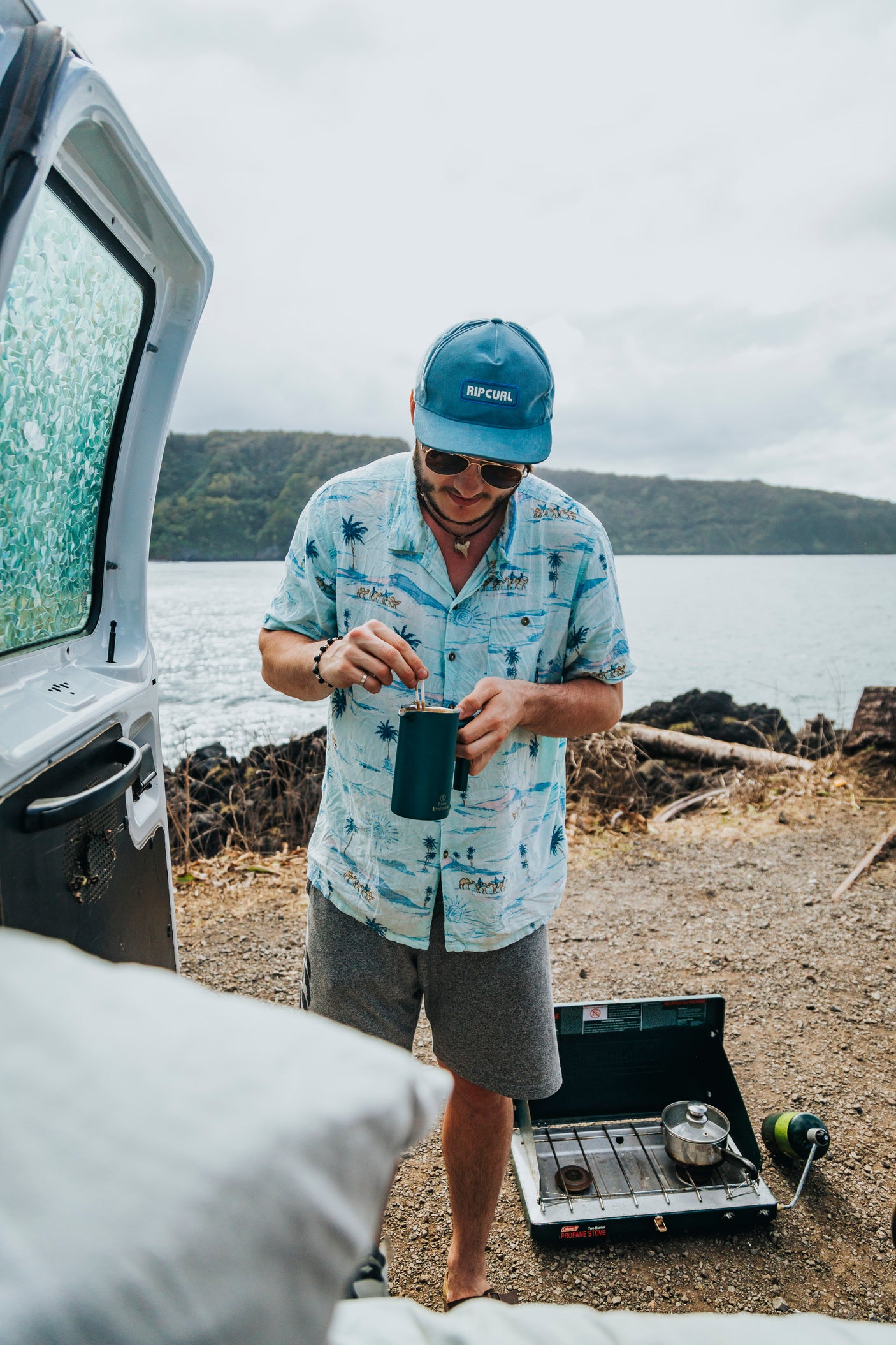 A man in a Rip Curl hat and Hawaiian shirt stirs coffee from an rv rental, cooking on a stove by the water and lush hills.