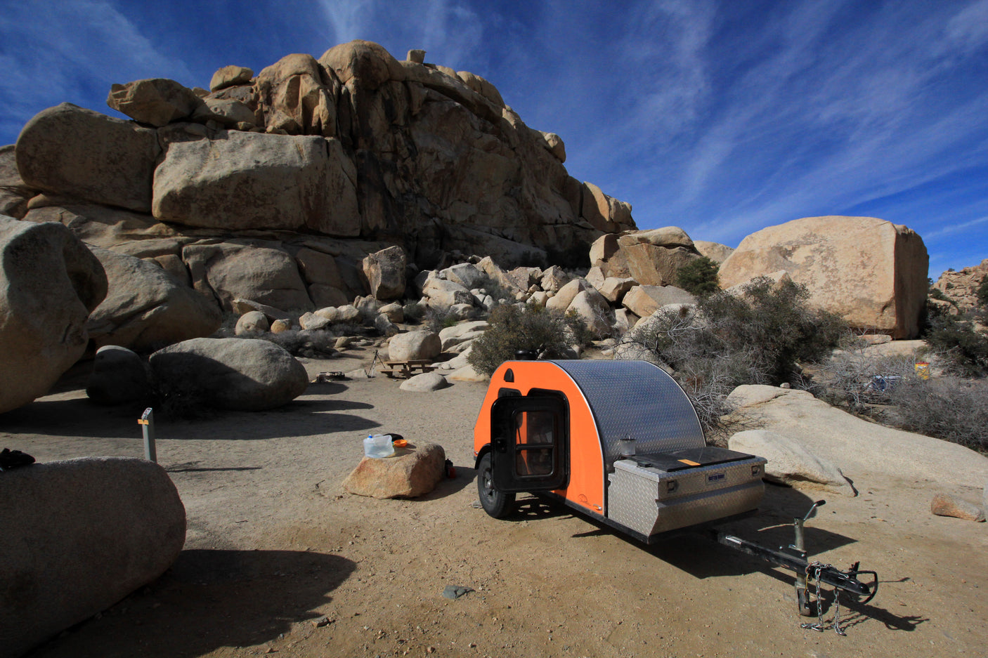 Orange teardrop RV rental camper at a desert campsite among large rocks under a blue sky, perfect for outdoor adventure.