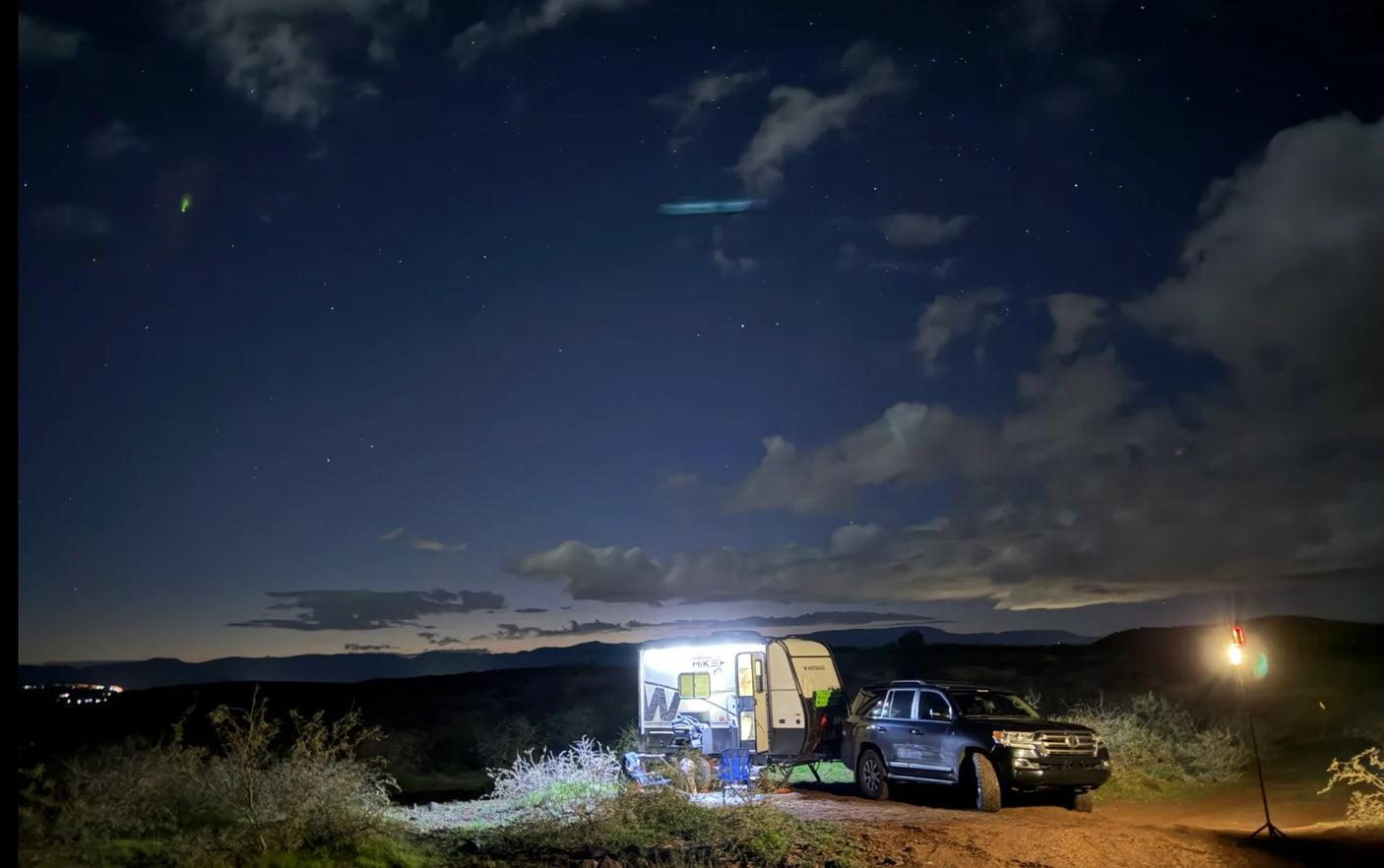 Night camping with an RV rental and SUV, lit under a starry sky. Green and blue lights are visible among the clouds.
