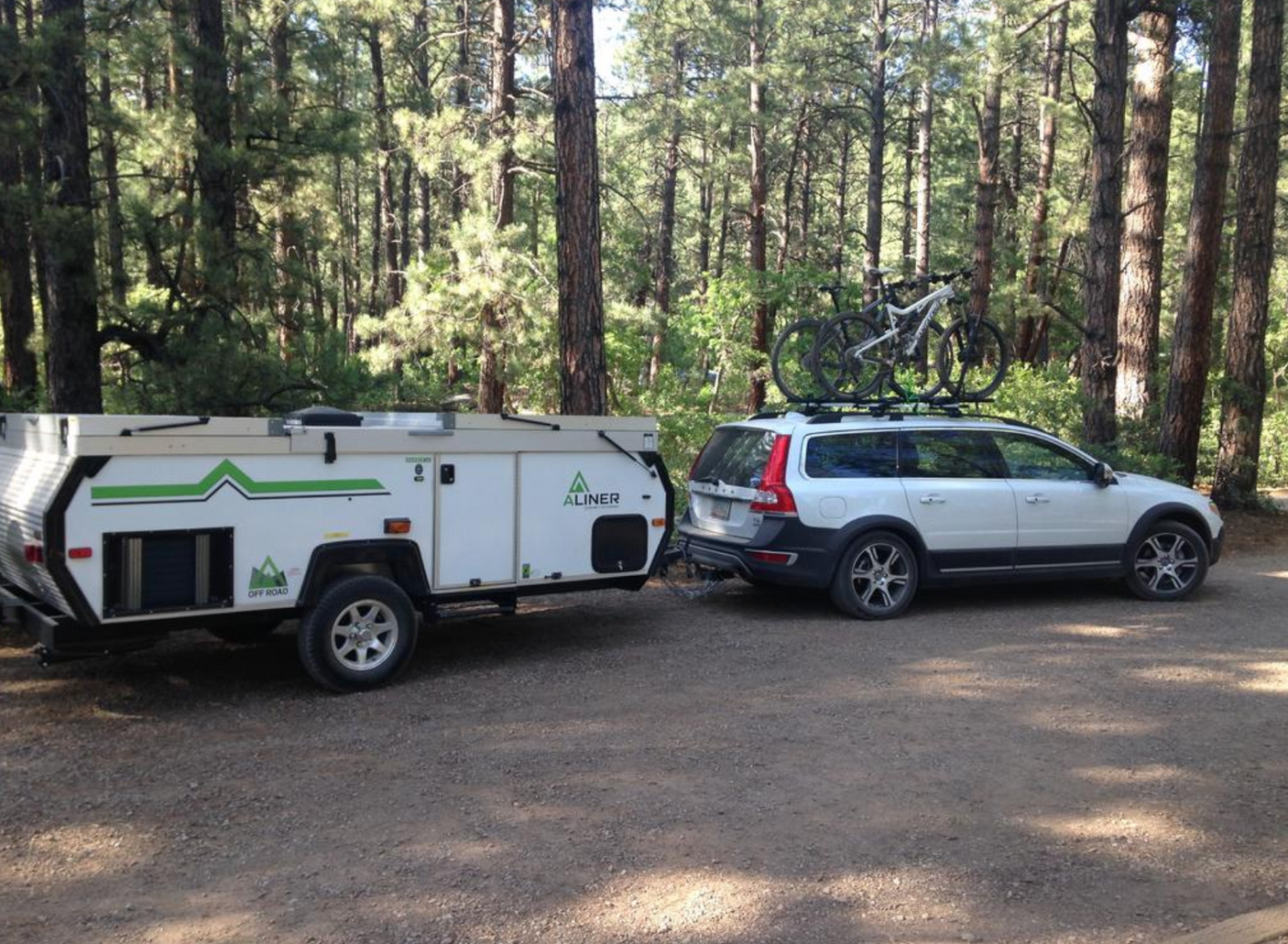 White Volvo XC70 towing an A-Liner pop-up camper with roof-mounted bikes at a forest campsite; great RV rental setup.