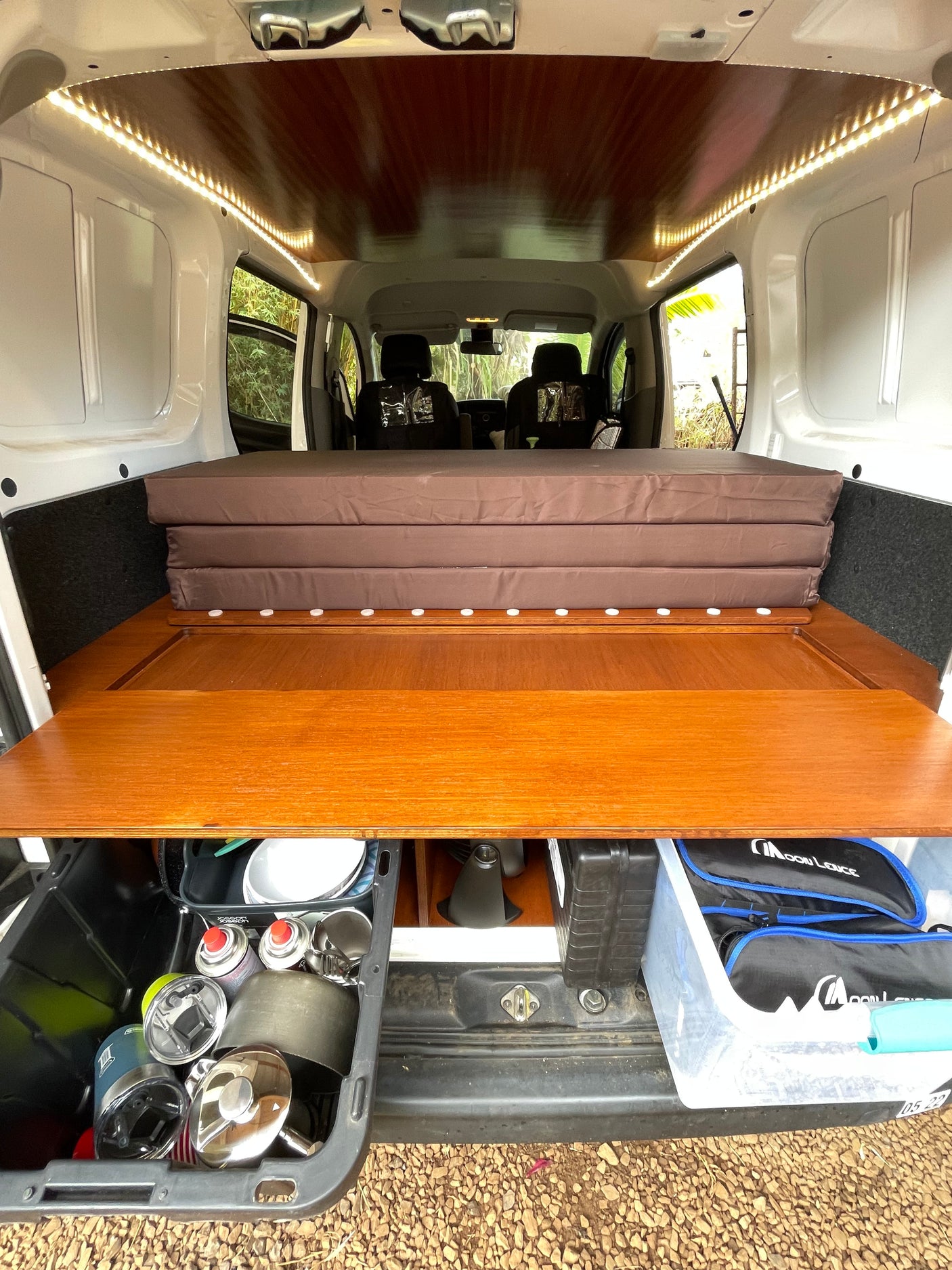 Inside a compact RV rental: a cozy wooden bed platform, LED lights, and pull-out storage with camp gear.