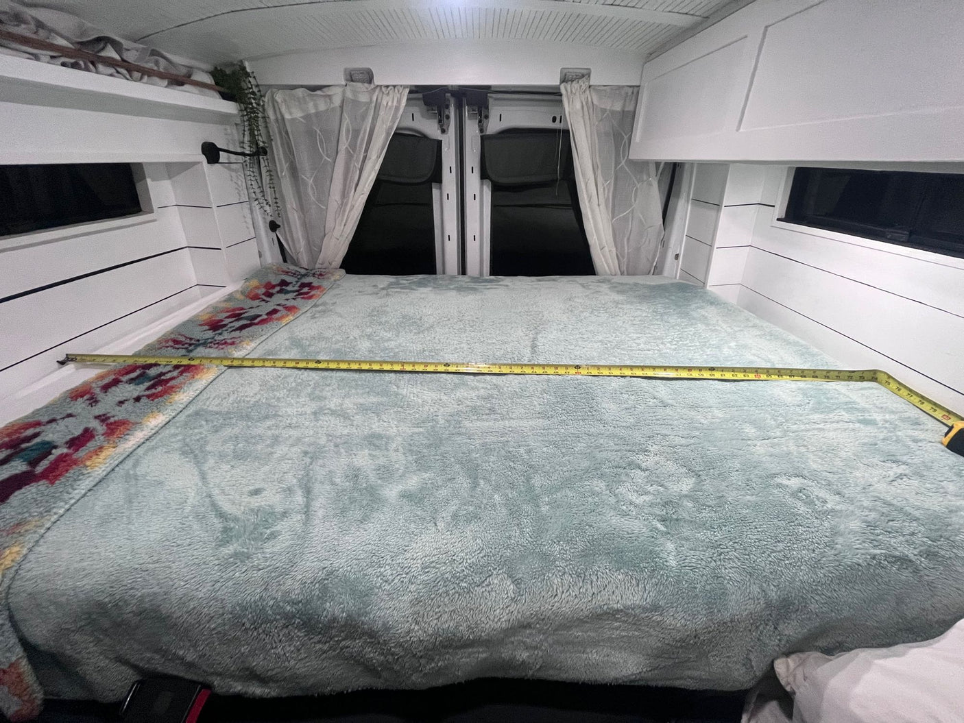 Inside an RV rental camper van: a cozy, light blue bed with a tape measure showing its width, nestled between white walls and curtains.