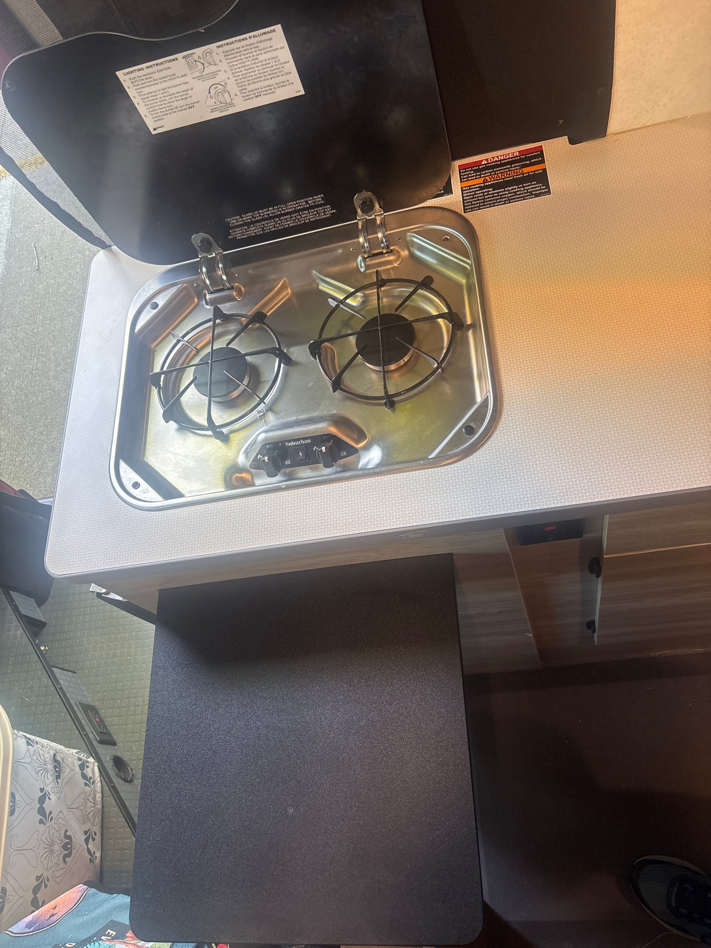 This RV rental features a 2-burner Suburban gas stove with safety warnings and lighting instructions visible on its open lid, ideal for a travel trailer kitchenette.