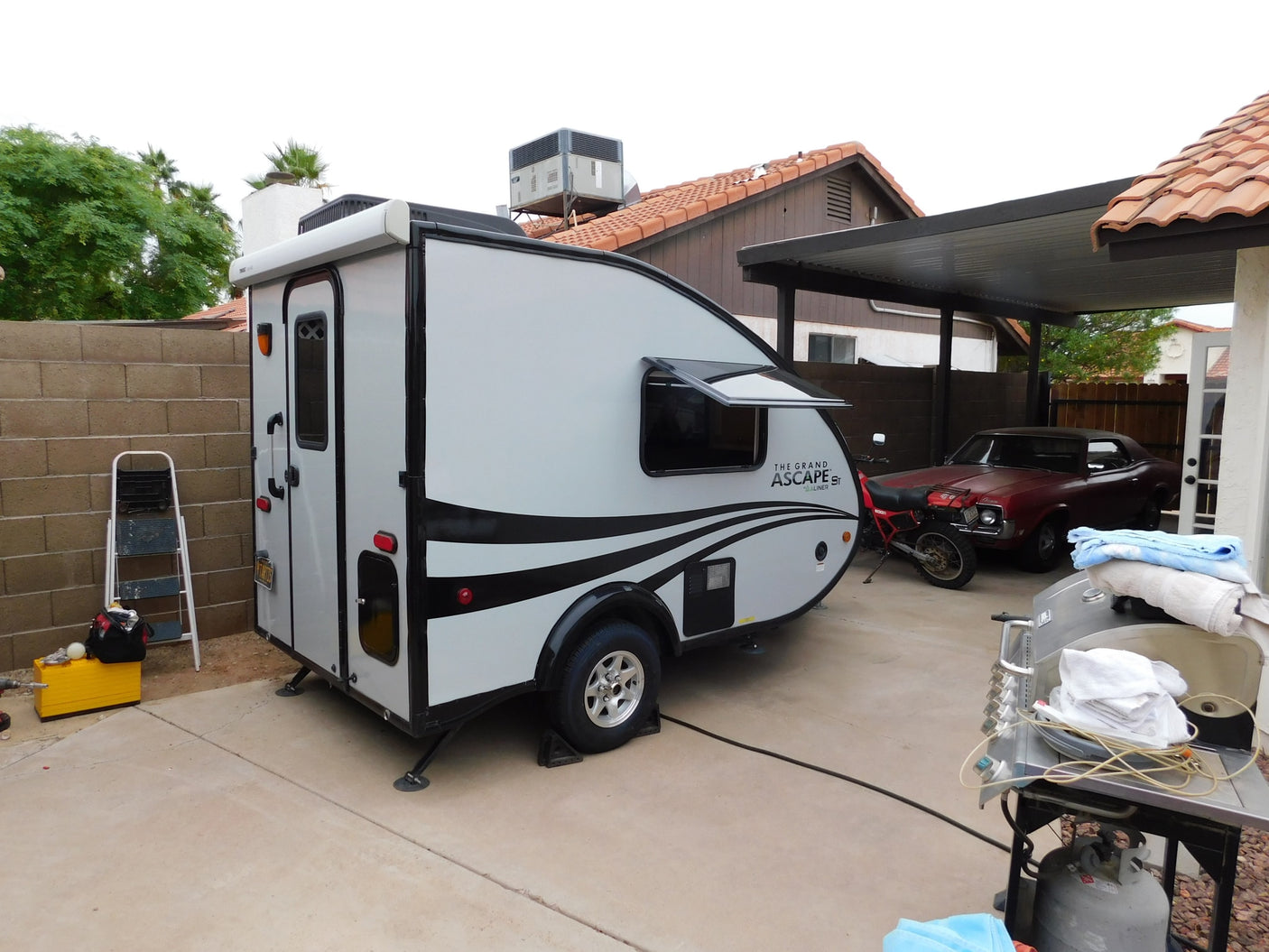 Grand ASCAPE ST RV rental travel trailer parked in a driveway, alongside a classic car and motorcycle.