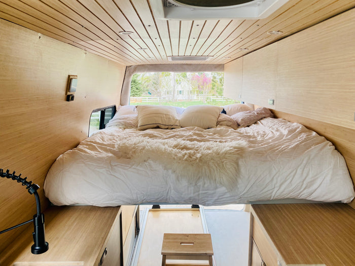 Interior of an RV rental camper van showing a large bed with white bedding, light wood walls, and a view outside.
