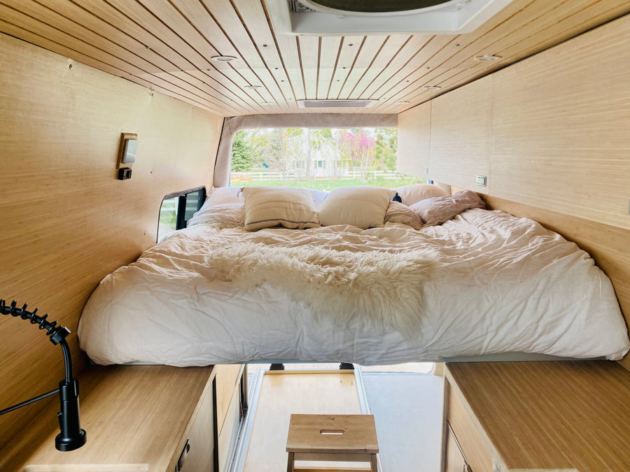 Interior of an RV rental camper van showing a large bed with white bedding, light wood walls, and a view outside.