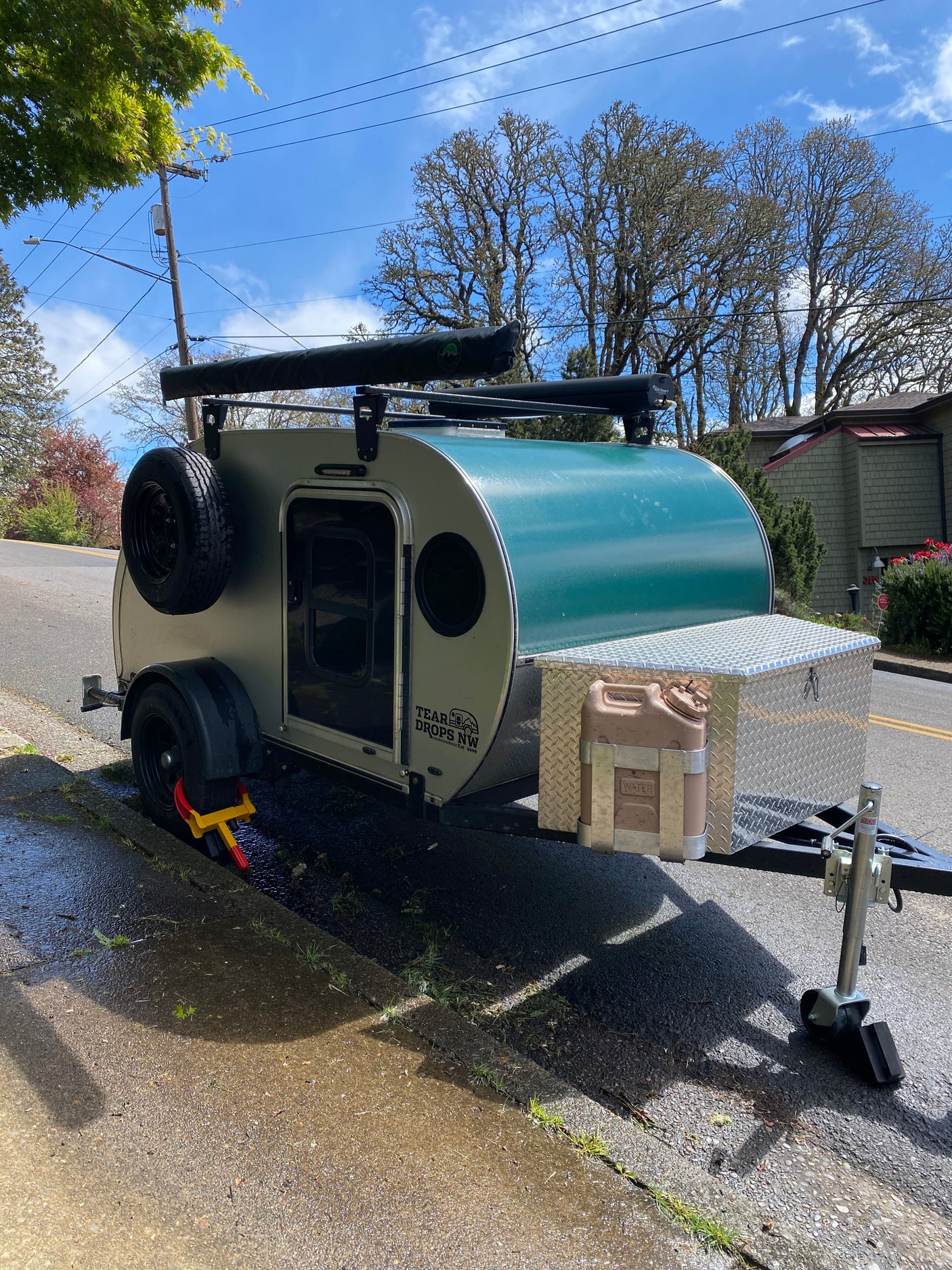 A green and grey teardrop **rv rental**, secured with a wheel lock, sits curbside ready for adventure.
