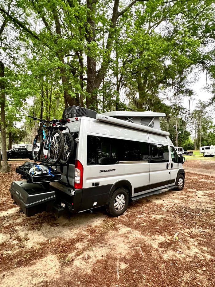 rests on gravel under a grand tree canopy.|Gray pop-top RV rental at a lakeside campground