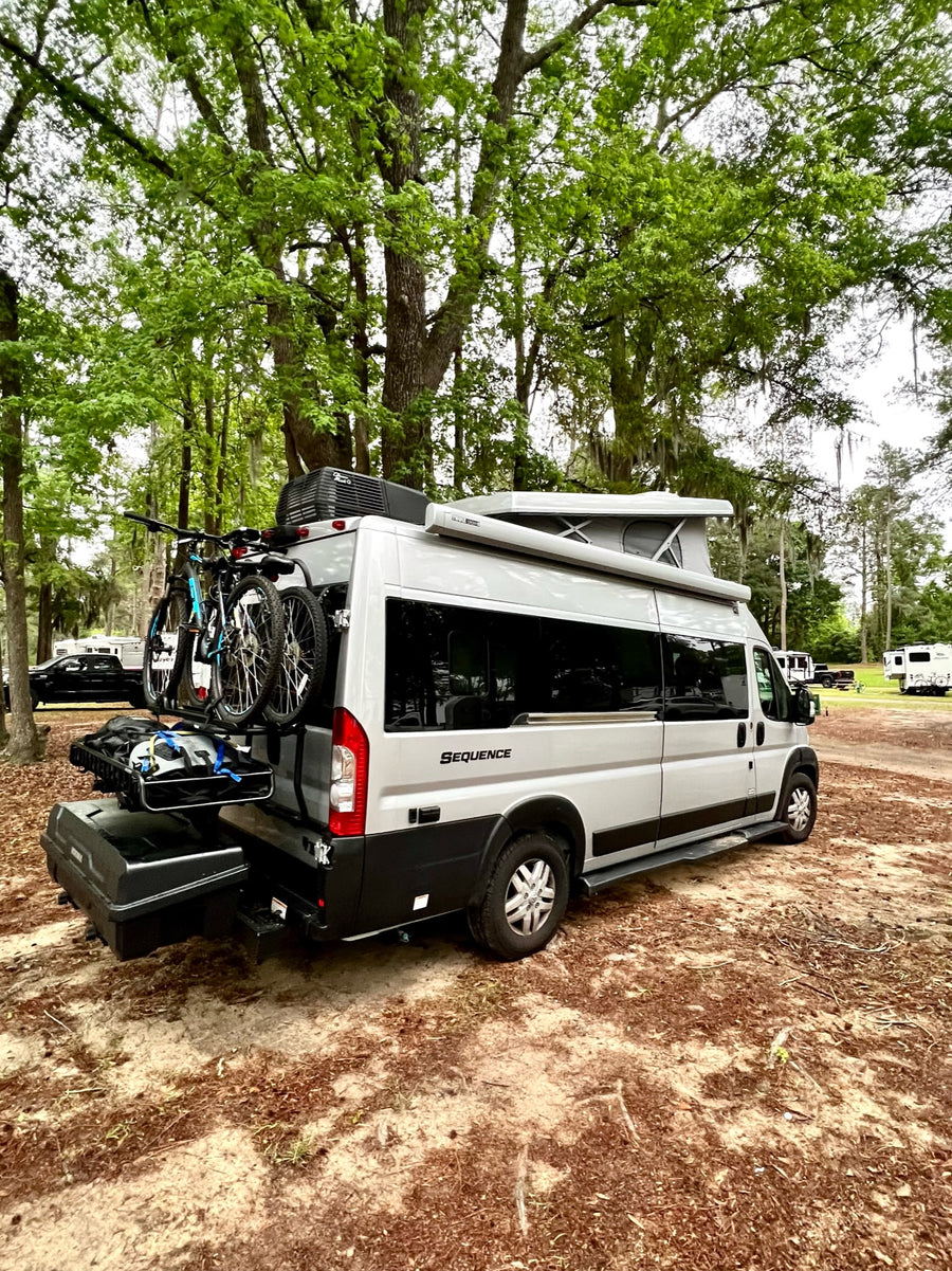 rests on gravel under a grand tree canopy.|Gray pop-top RV rental at a lakeside campground