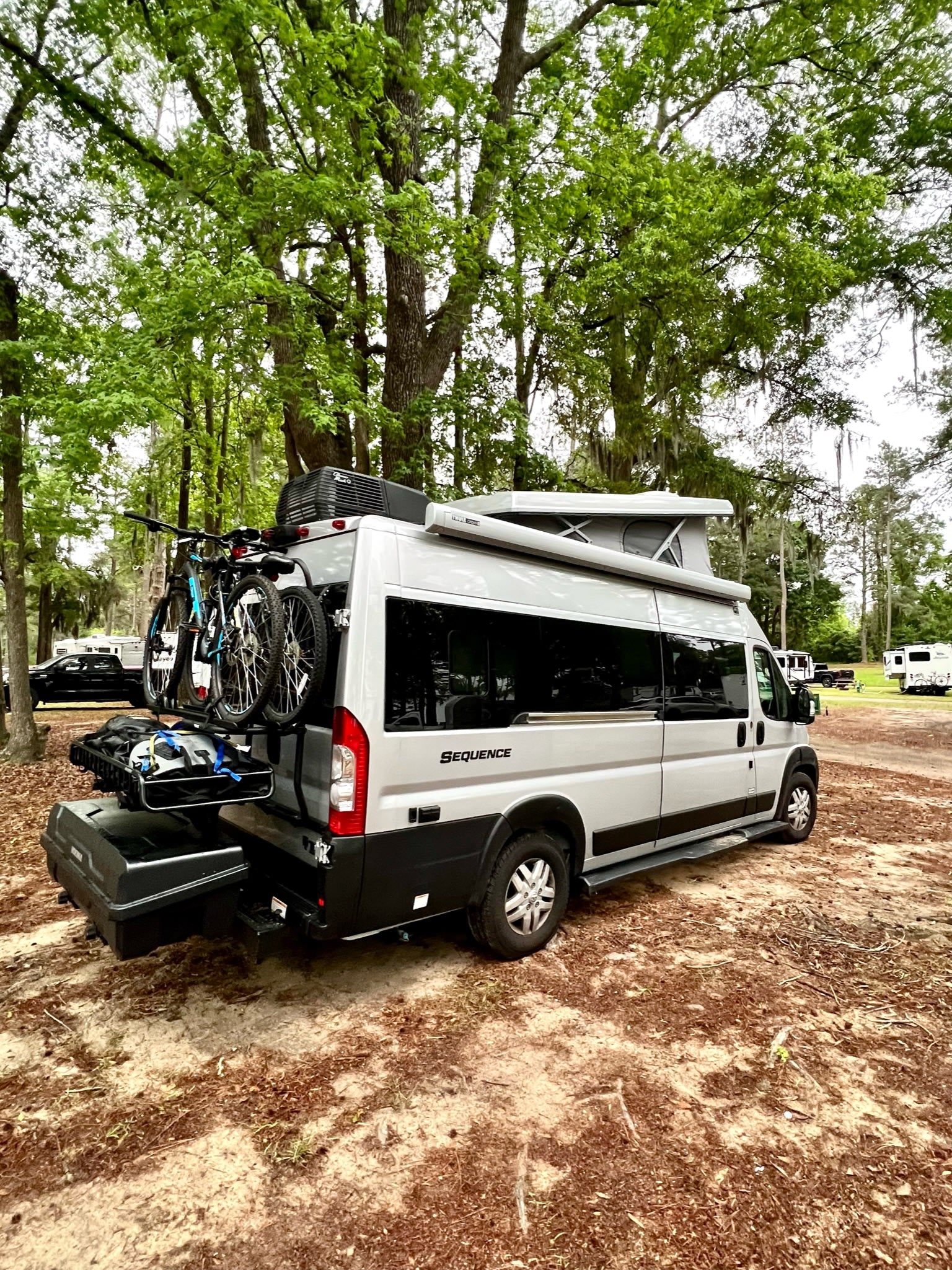 rests on gravel under a grand tree canopy.|Gray pop-top RV rental at a lakeside campground