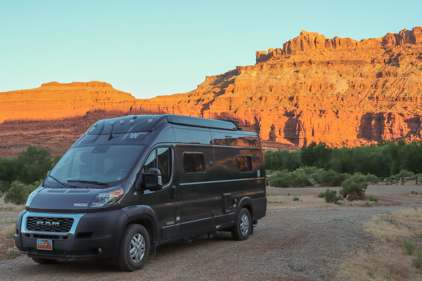 Dark gray Winnebago Solis RV rental on a dirt road, bathed in warm light from striking red rock mountains and green trees.