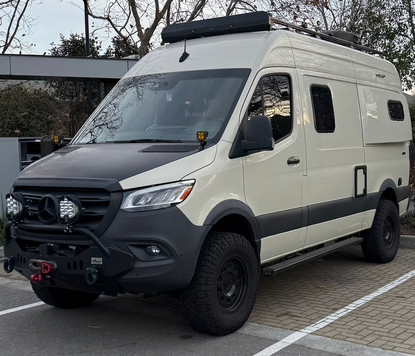 An off-white Mercedes Sprinter camper van, modified for off-road travel with a roof rack and rugged tires, ready for rv rental.