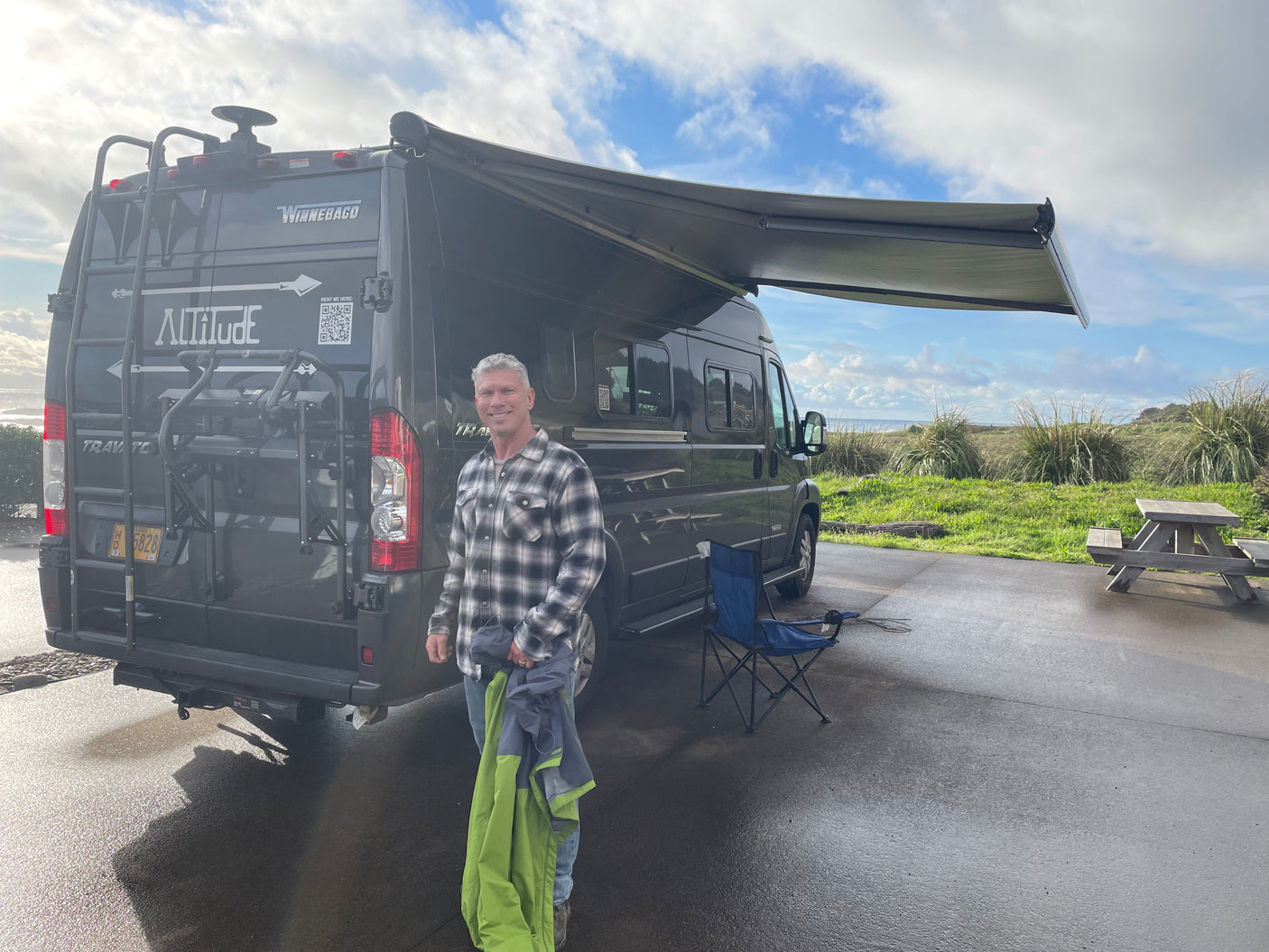 Man smiles by his dark gray Winnebago rv rental van, parked with awning out at a scenic beachside campsite.