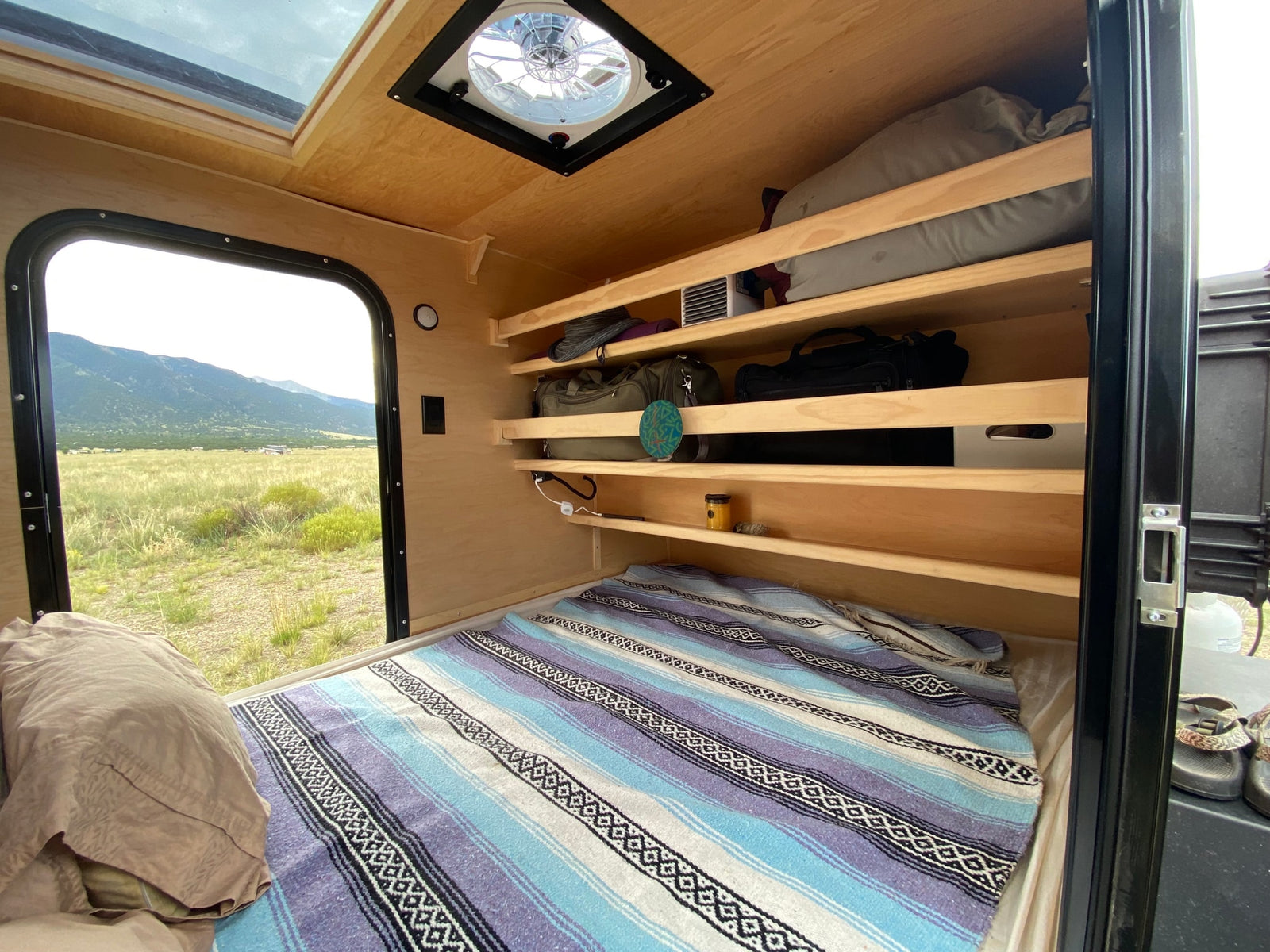 Experience comfort in this RV rental's interior: bed, organized shelves, ceiling vent, & scenic window to mountains.