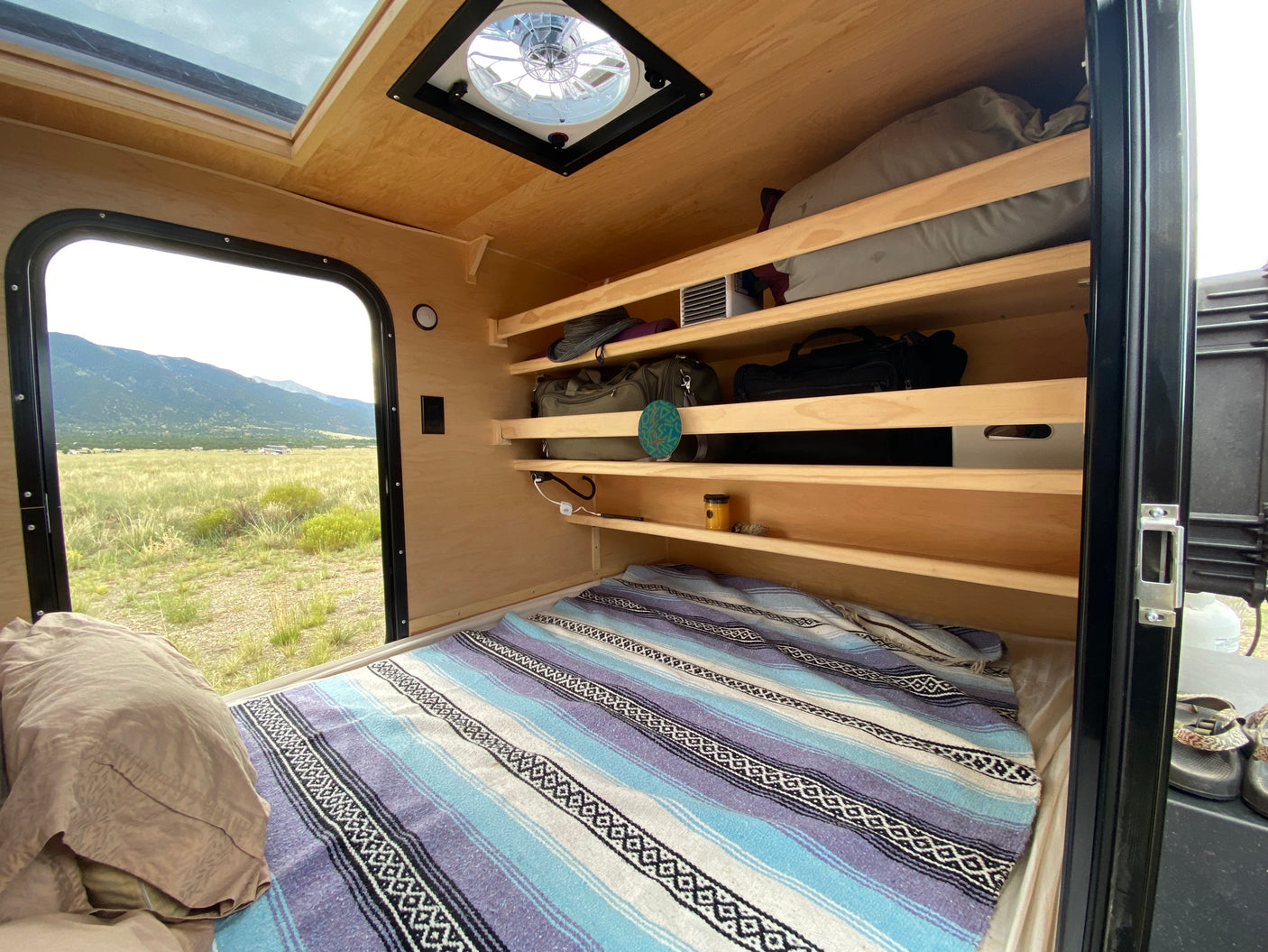 Experience comfort in this RV rental's interior: bed, organized shelves, ceiling vent, & scenic window to mountains.