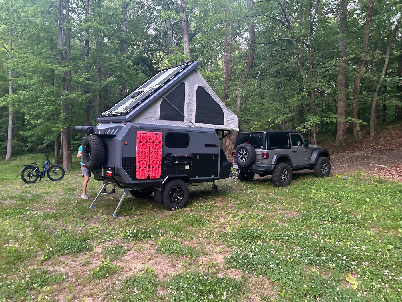 RV rental: Grey off-road camper trailer with rooftop tent & solar panels, hitched to a Jeep in a wooded campsite.
