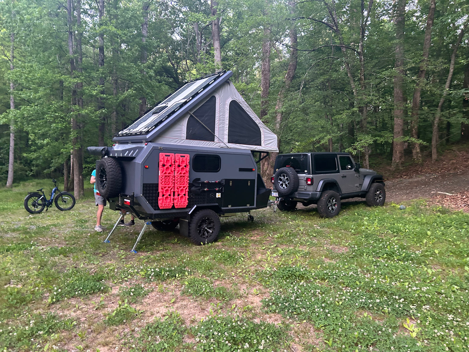 RV rental: Grey off-road camper trailer with rooftop tent & solar panels, hitched to a Jeep in a wooded campsite.