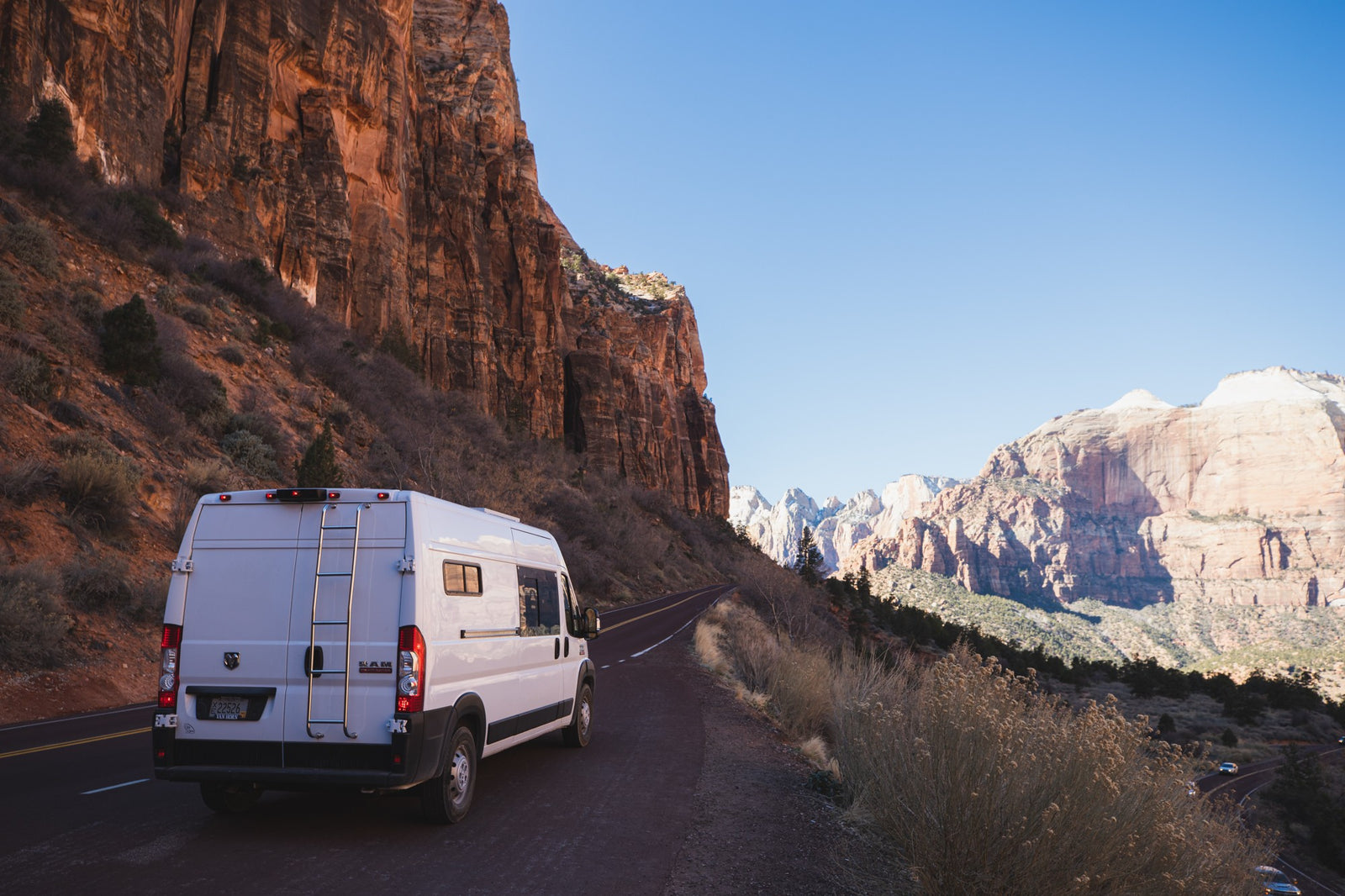 This RV rental van is parked on a winding road amidst the towering red cliffs of Zion National Park. Clear blue sky.