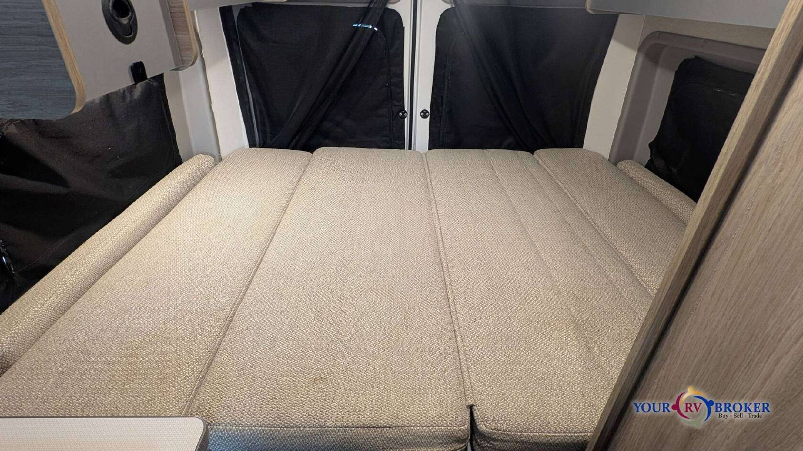 This RV rental features a folded-out bed with light-colored cushions and black privacy curtains, inside a cozy camper.