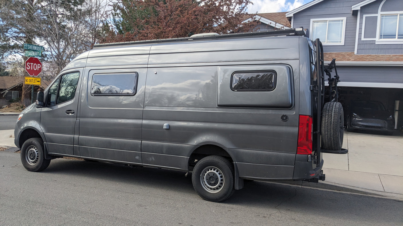 This custom gray Mercedes Sprinter camper van is perfect for rv rental trips. It has a roof rack, rear gear carrier, and distinctive windows.