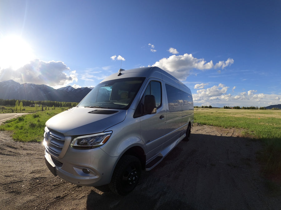 Experience nature with this silver RV rental. Parked on a dirt path, with sunny mountains and green fields.
