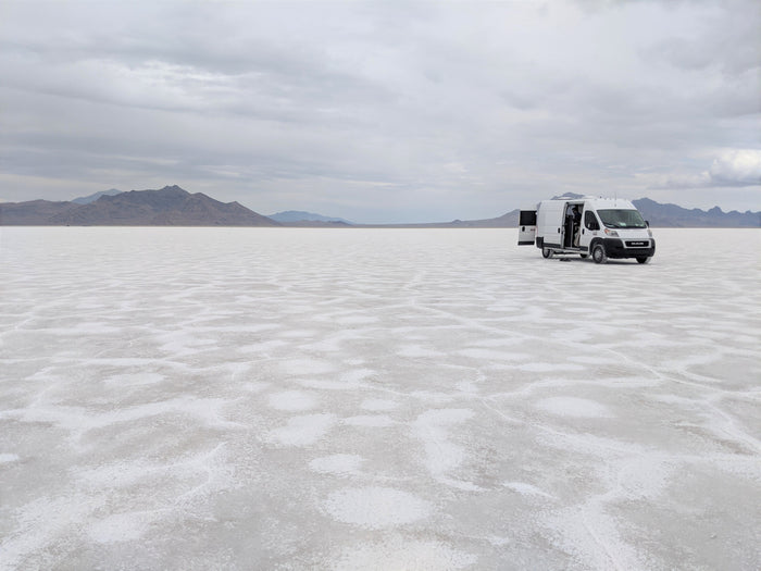 White RV rental van on vast Bonneville Salt Flats with open doors, mountains under cloudy sky.
