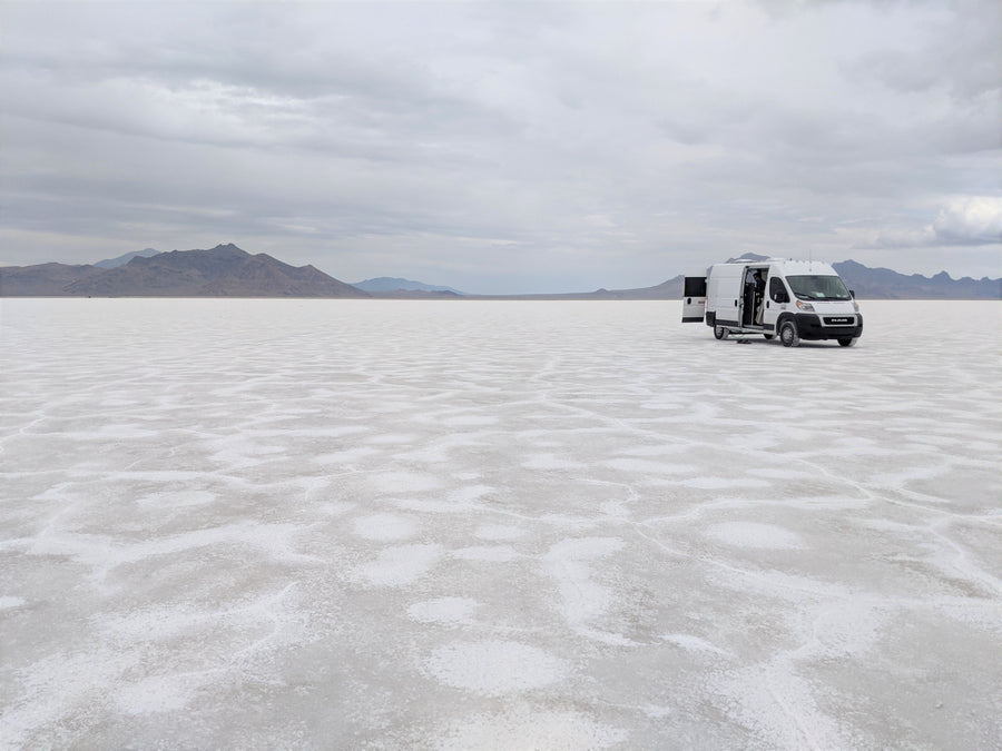 White RV rental van on vast Bonneville Salt Flats with open doors, mountains under cloudy sky.