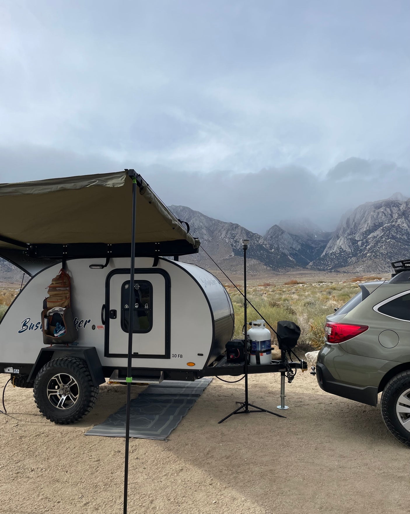 This RV rental, a teardrop trailer with an awning, is set up for camping in front of cloudy mountains, hitched to a Subaru.
