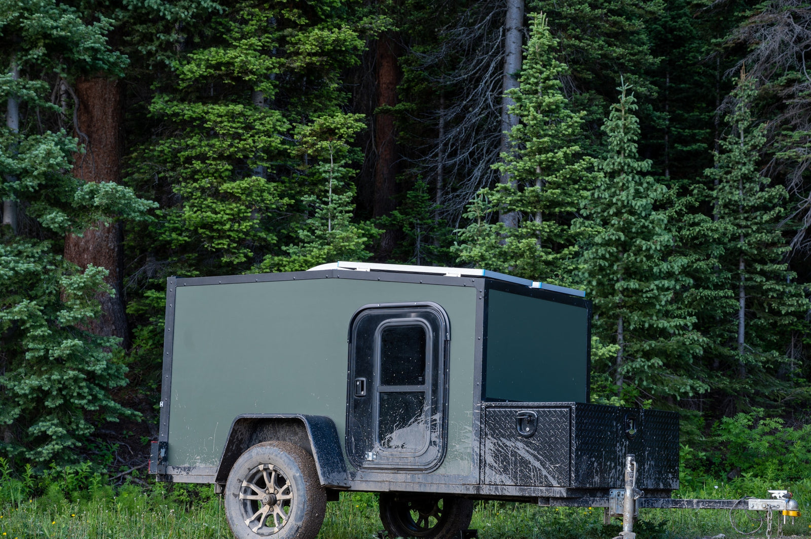 Mud-splattered green off-road RV rental trailer nestled in a dense forest, ready for adventure.