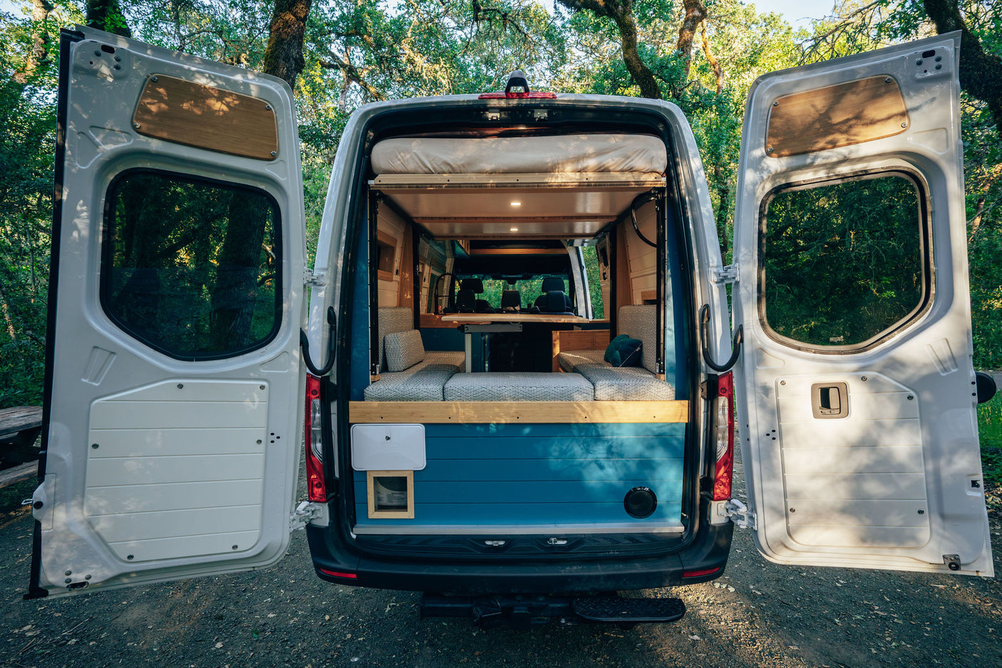 Open doors of an RV rental camper van showcase its cozy, converted interior with a bed and seating, parked in nature.