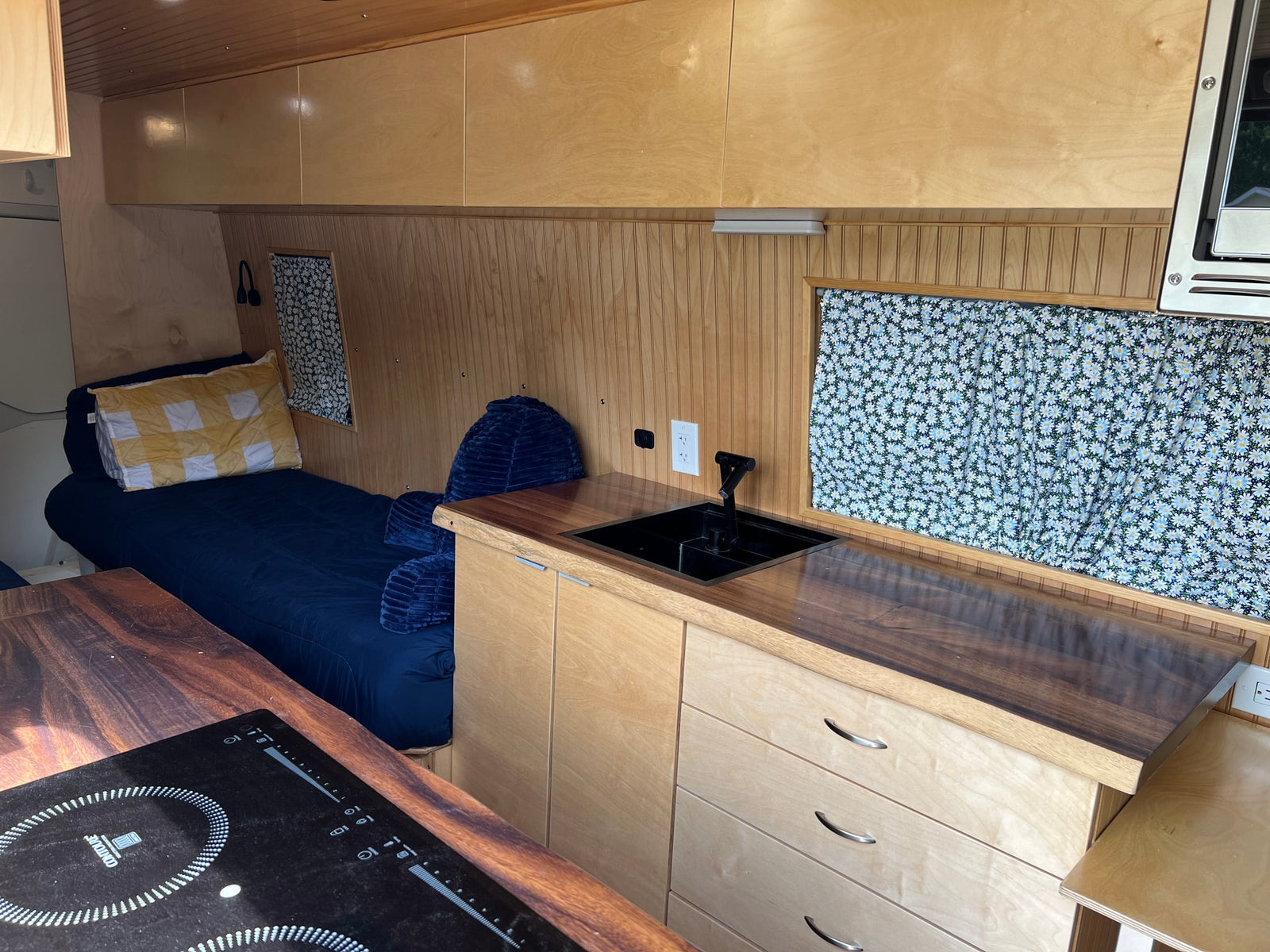 Cozy RV rental interior with a wood-accented kitchen, comfortable bed, and charming daisy window curtains.