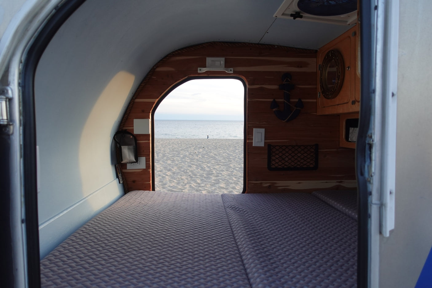 Cozy rv rental interior with bed and wood details, looking out to a serene sandy beach and calm ocean.