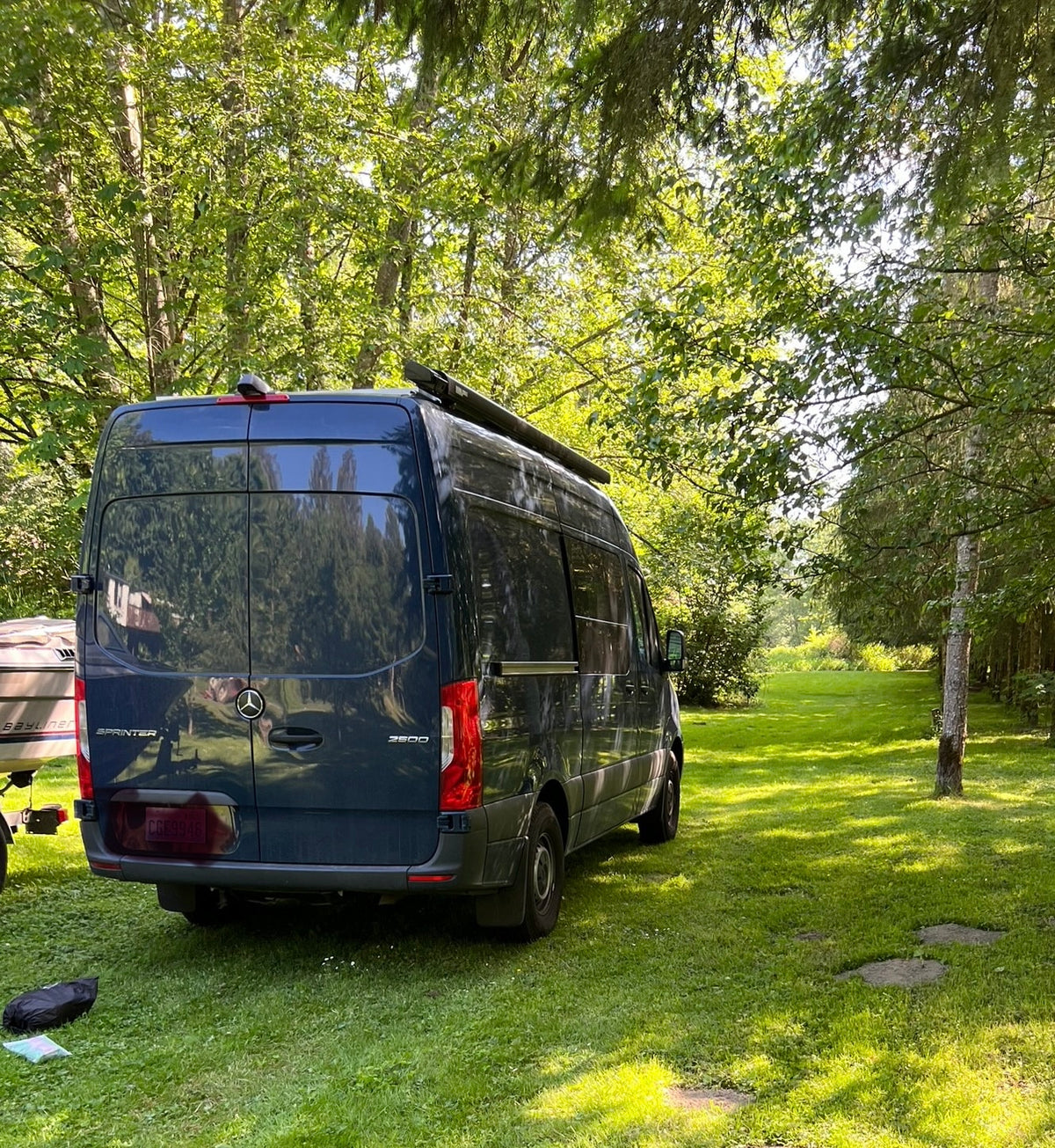 This dark blue Mercedes Sprinter rv rental is parked in a sunny, wooded area. Perfect for camping and van life outdoors. — photo 1
