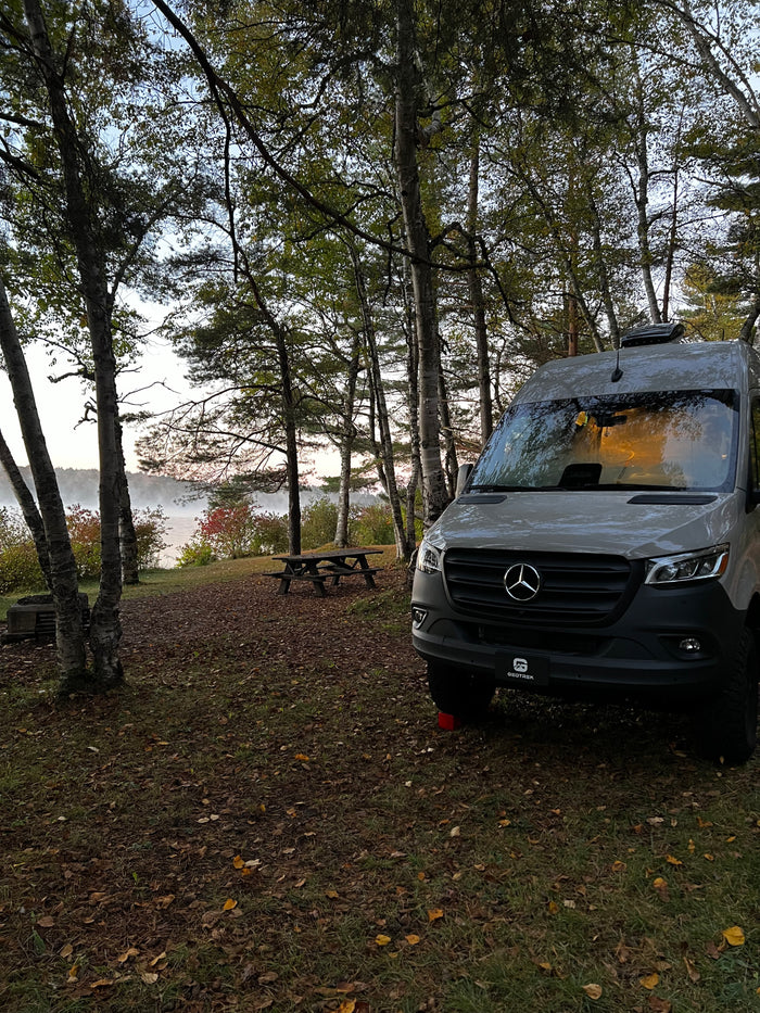 RV rental camper van parked by a misty lake, surrounded by autumn trees and a picnic table at a scenic campsite.
