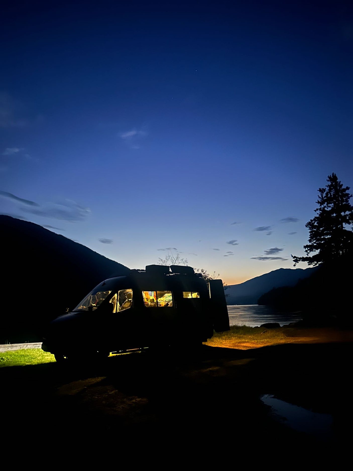 Cozy RV rental parked by a mountain lake at dusk. Warm lights glow inside the campervan.