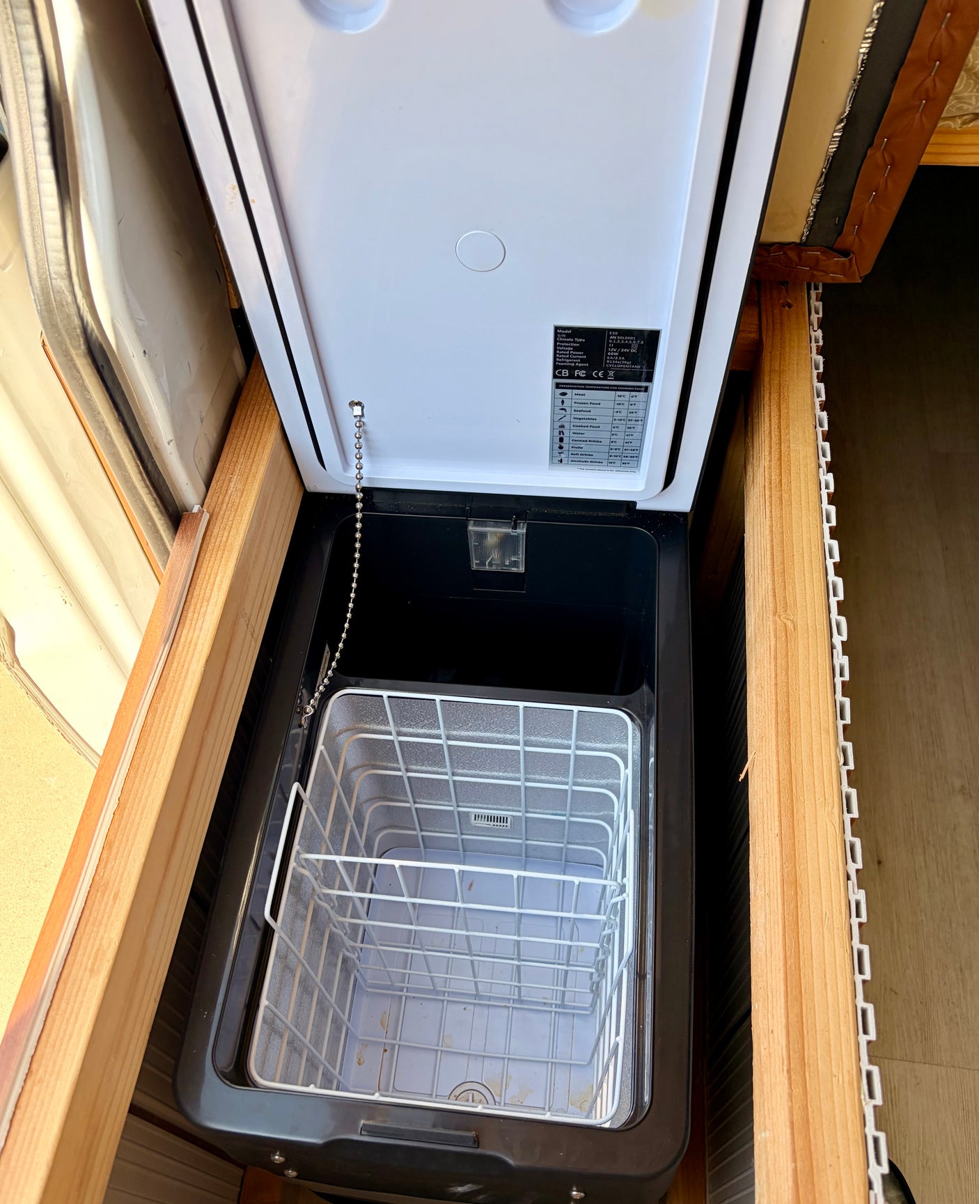 Inside an rv rental, a compact portable fridge/freezer is open, revealing its wire basket and essential specs on the lid for mobile travel.