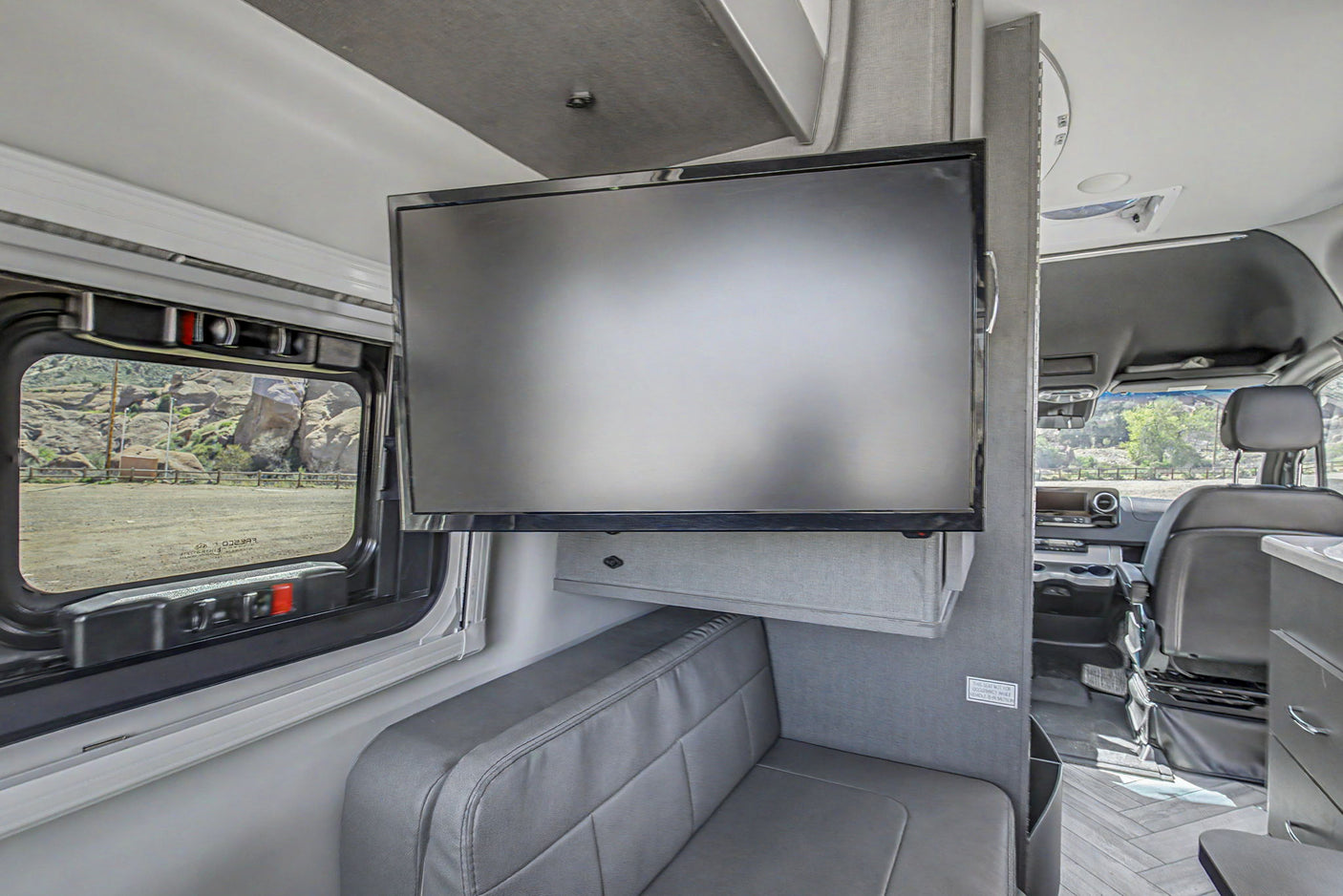 Inside a modern RV rental: TV, gray bench seat, and a window framing a rocky landscape. Comfort for your mobile adventures.