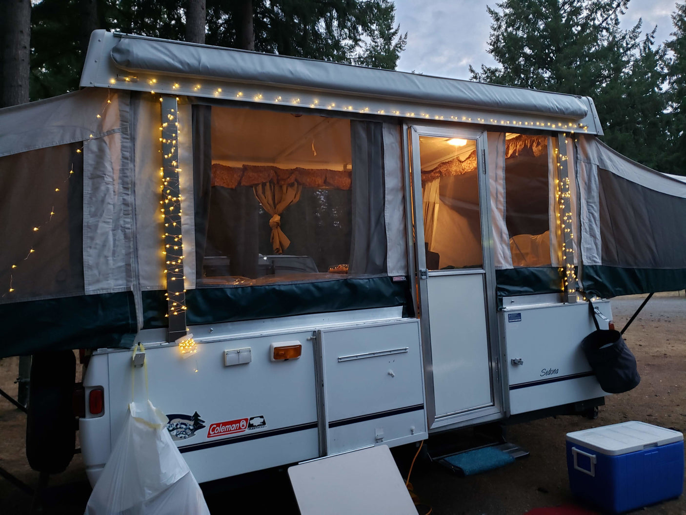 An inviting Coleman Sedona pop-up camper rv rental with warm string lights set up at a dusky campsite.|RV rental pop-up camper & SUV at a sunny campsite with a fire pit and concrete picnic table.|Inside this cozy RV rental: a compact kitchen with sink and stovetop