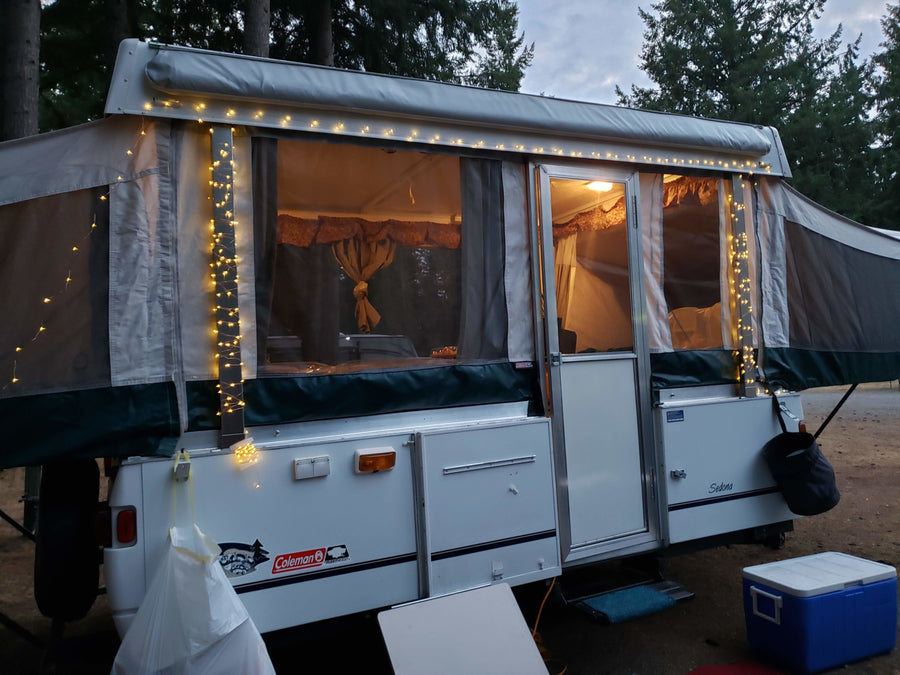 An inviting Coleman Sedona pop-up camper rv rental with warm string lights set up at a dusky campsite.|RV rental pop-up camper & SUV at a sunny campsite with a fire pit and concrete picnic table.|Inside this cozy RV rental: a compact kitchen with sink and stovetop
