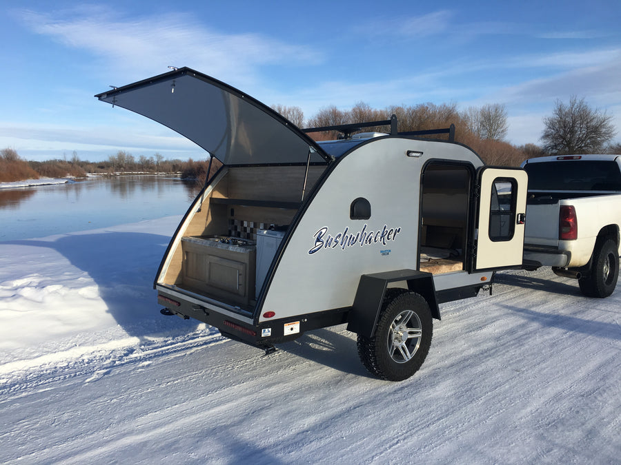 Bushwhacker teardrop RV rental on a snowy riverside, showing its outdoor kitchen & interior. Perfect for winter camping.
