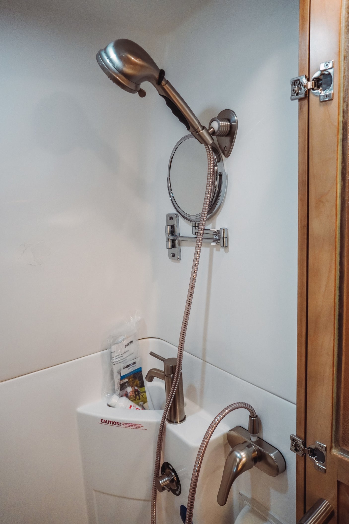 Inside a compact RV rental bathroom, showing a shower, sink, adjustable mirror, and various controls.