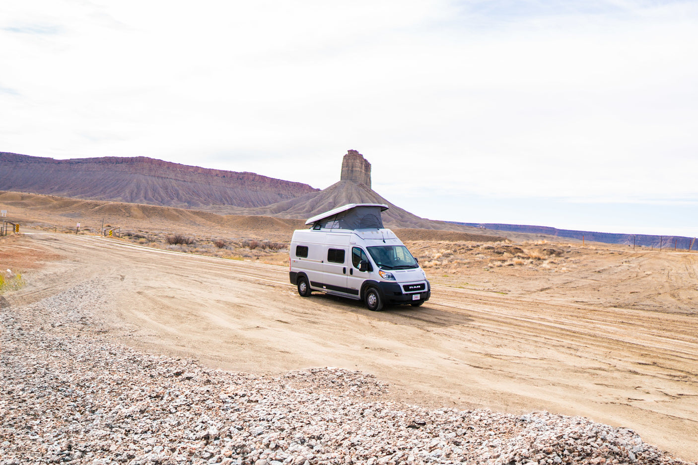 White pop-top RV rental on a dirt road, amidst stunning desert mountains and unique rock formations. Ready for adventure.
