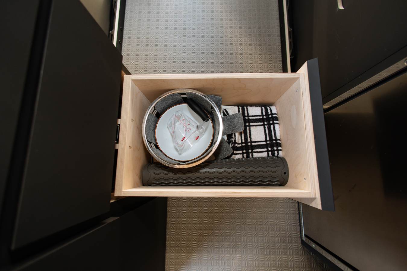 Organized wooden drawer in an RV rental, showing kitchen essentials: pot, measuring cup, towel, and silicone mat.