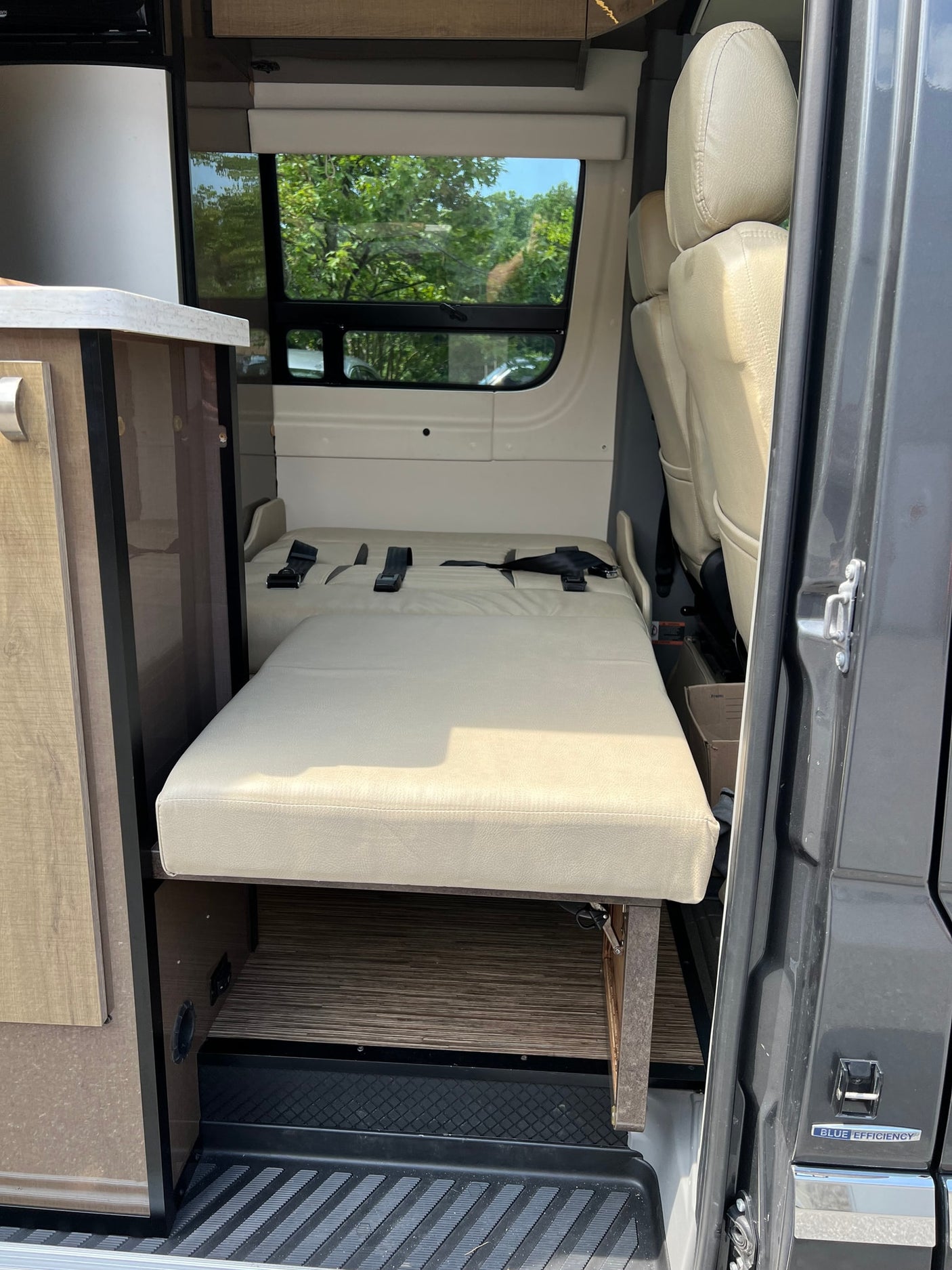 parked on a driveway surrounded by trees.|Front view of a modern gray Mercedes-Benz Sprinter van
