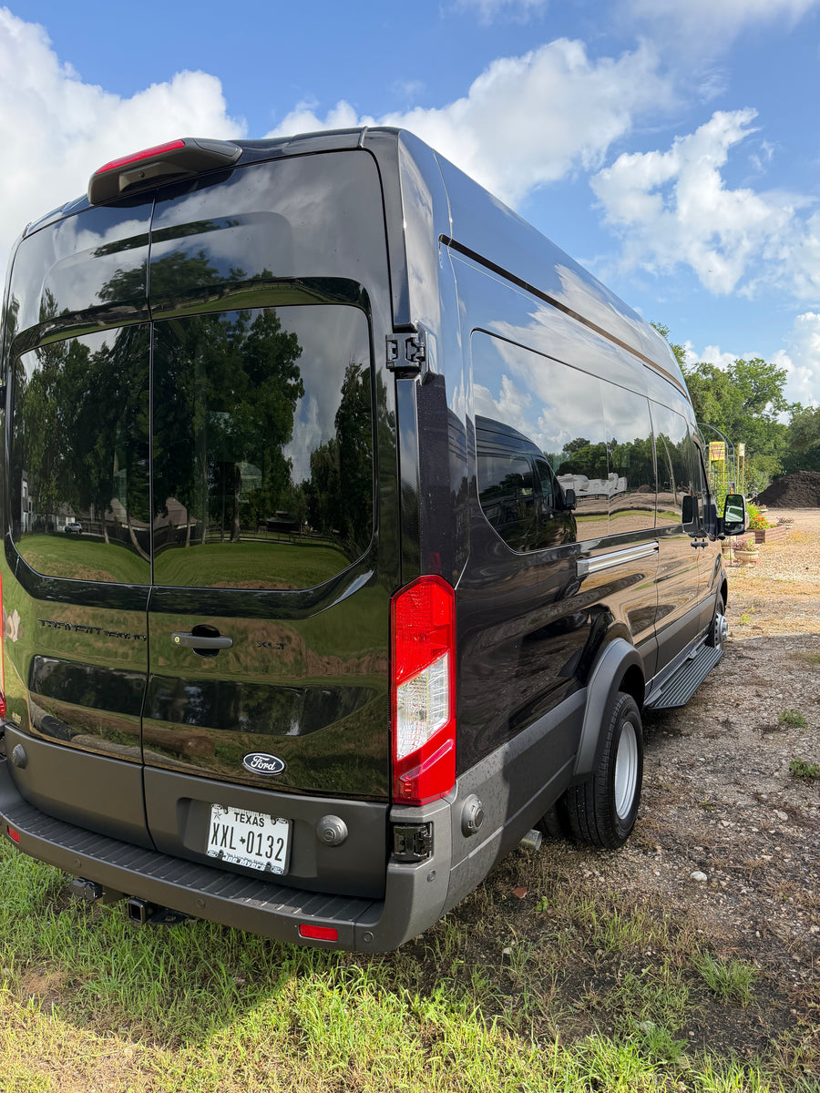 Black Ford Transit van for RV rental, detailed from the rear, reflecting sky & trees on its sleek surface.